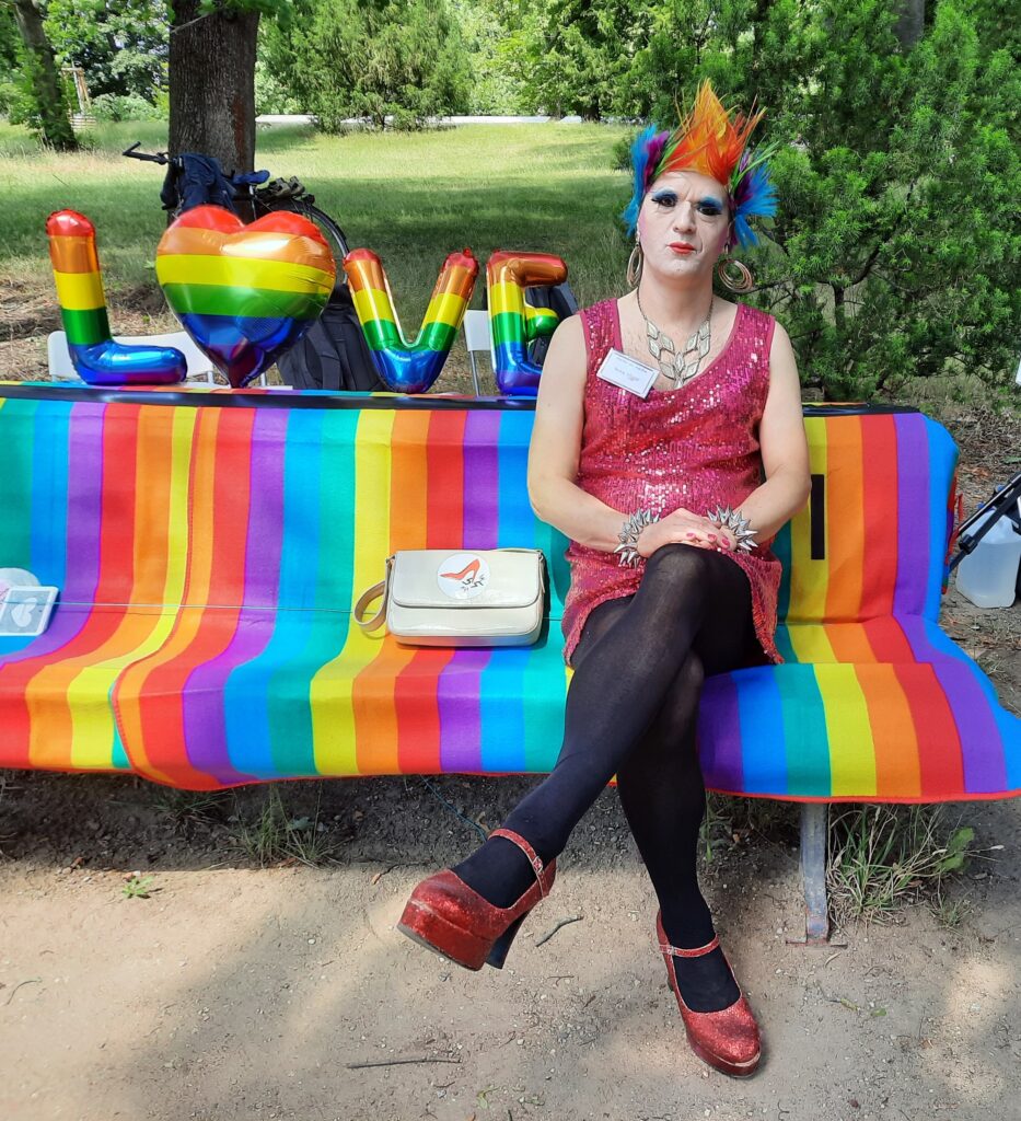 Drag Queen in Paillettenkleid und mit bunter stacheliger Kurzhaar-Perrücke sitzt im Park auf einer Bank mit Regenbogendecke.